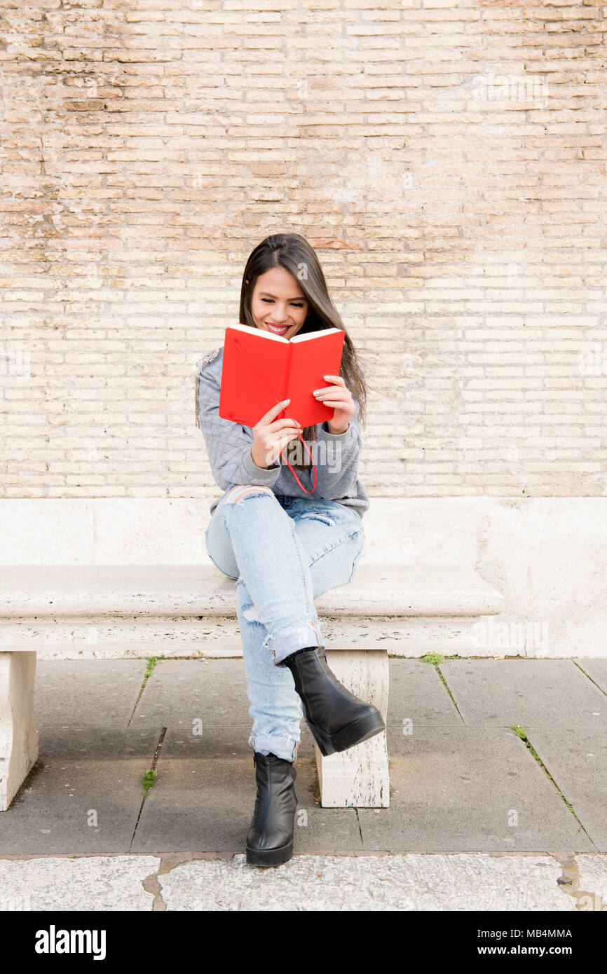 Young latina woman sitting on old marble bench smiling reading red book in hands Stock Photo