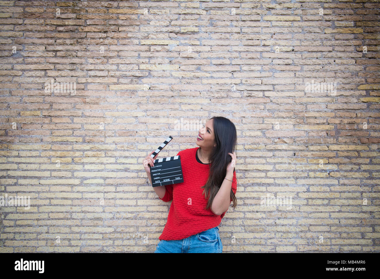 Young pretty latina woman holding movie clapper board with brick wall in background looking up Stock Photo