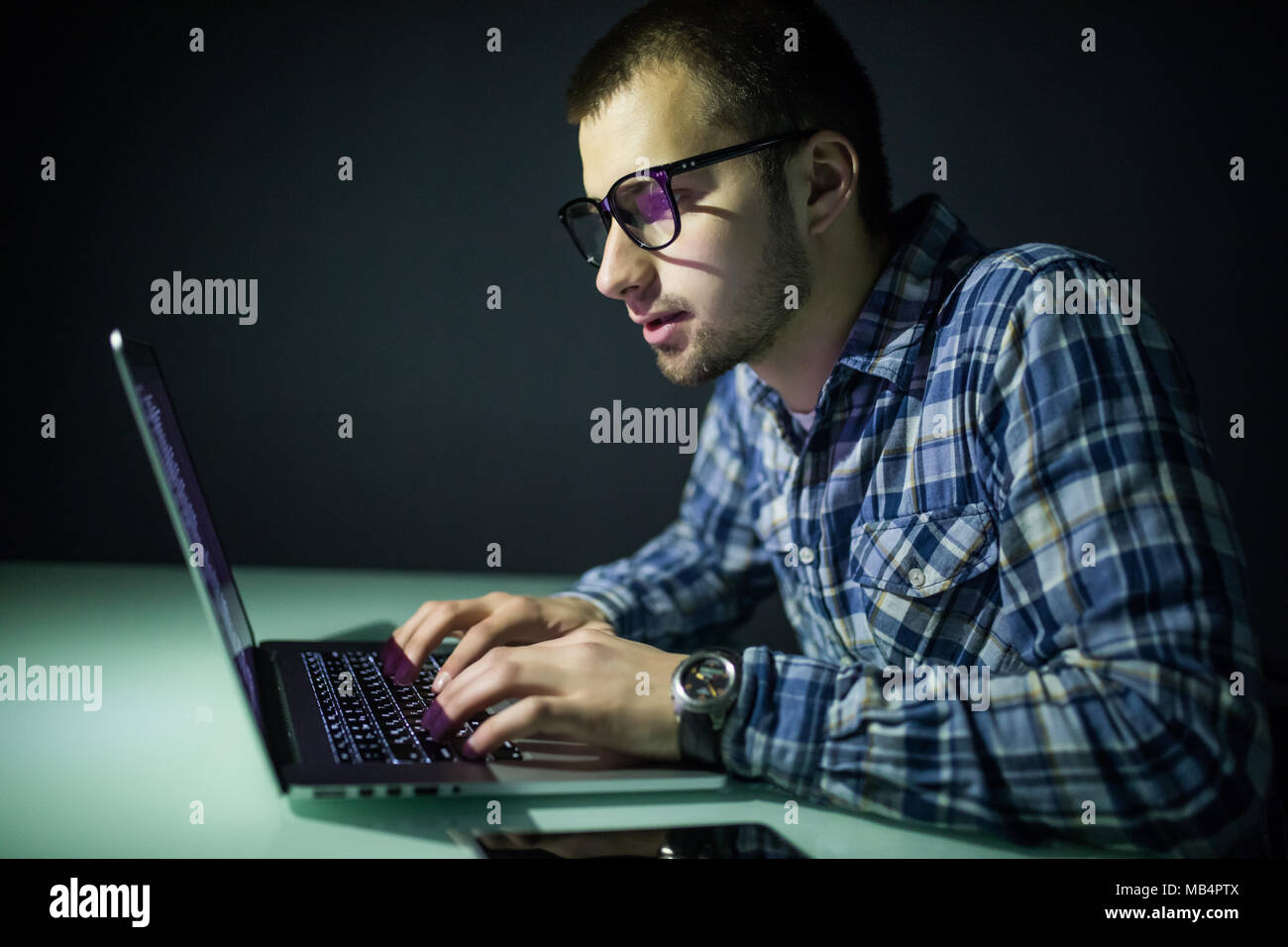 Concentrated young hacker using computer and stealing information in dark room Stock Photo