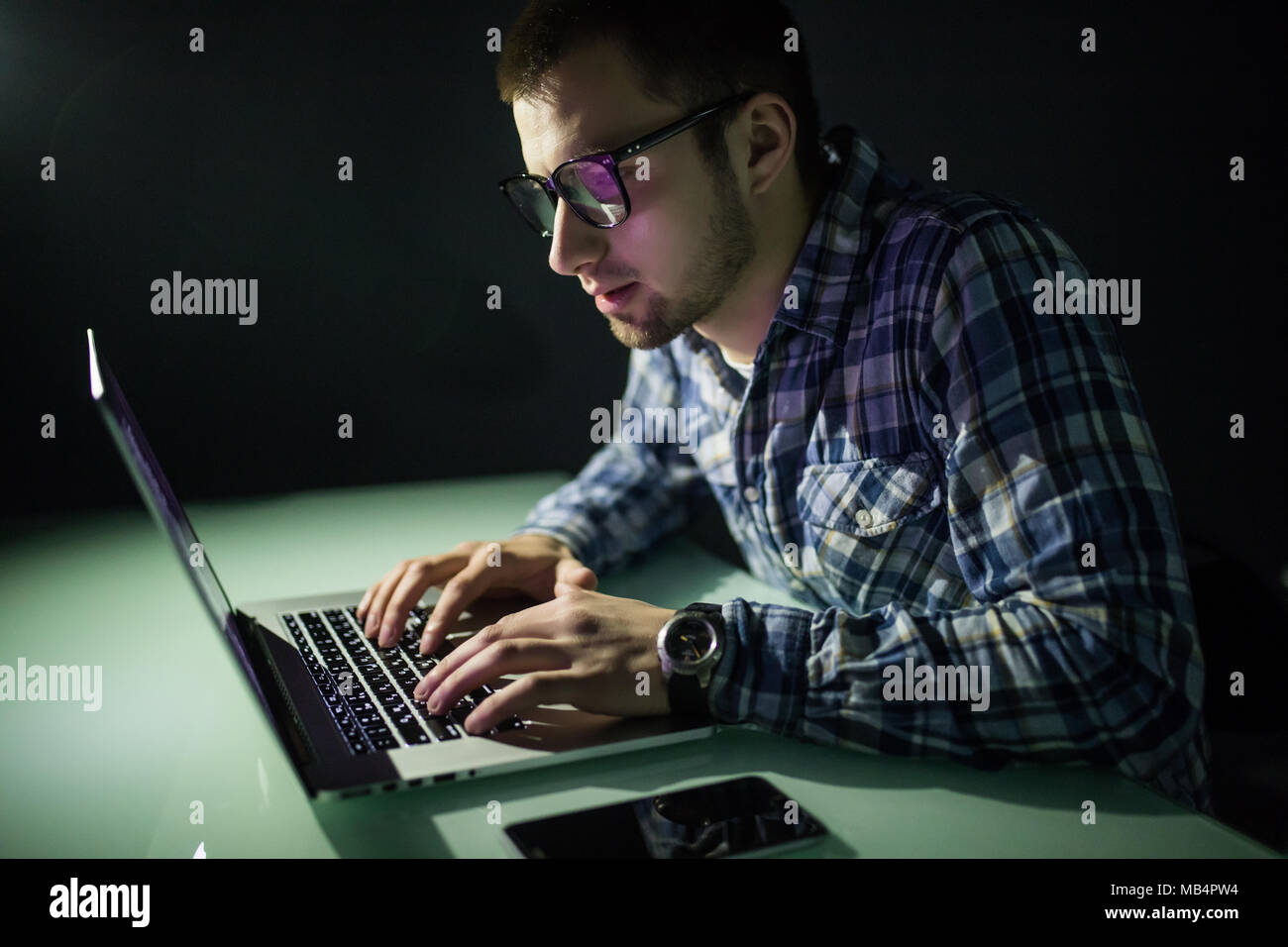 Concentrated young hacker using computer and stealing information in dark room Stock Photo