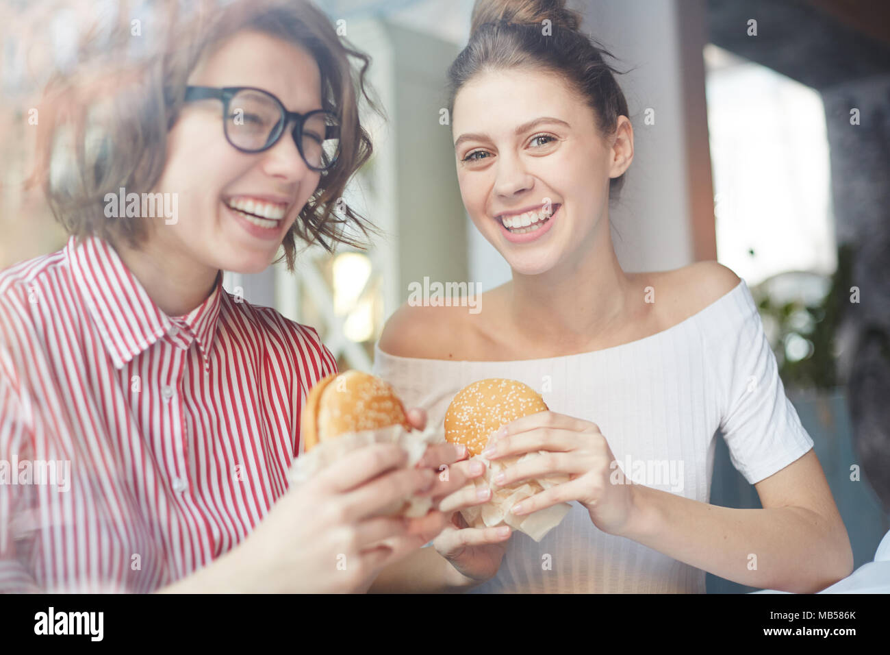 Two happy girls with appetizing and fat cheeseburgers having fun and enjoying time in fastfood cafe Stock Photo