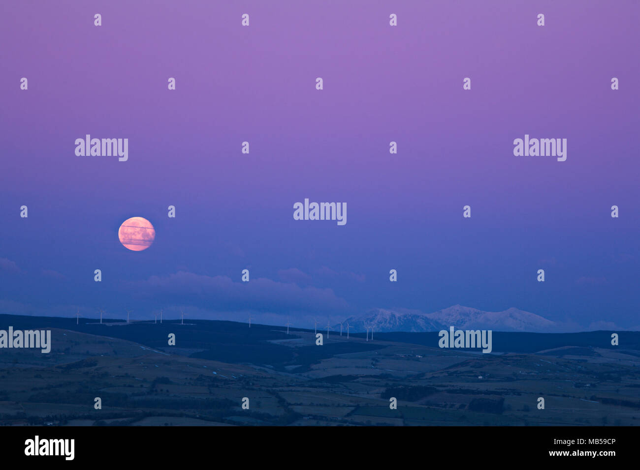 Full moon setting into earth shadow over snow capped mountains and wind turbines, North Wales. as seen from Moel Famau Stock Photo
