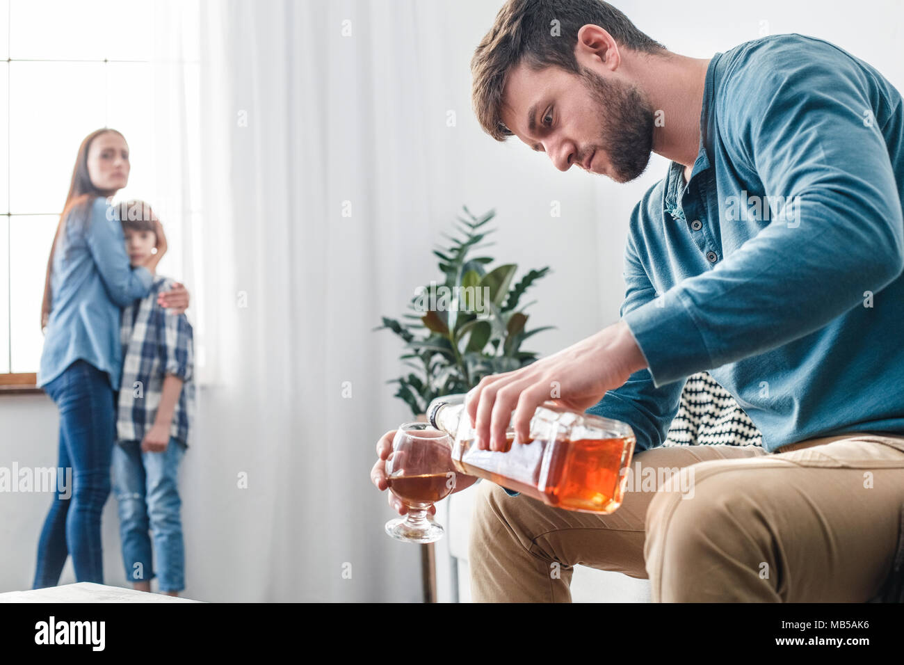 Mother father and son social problems alcoholism man pouring whiskey into glass while wife with kid standing near window scared Stock Photo