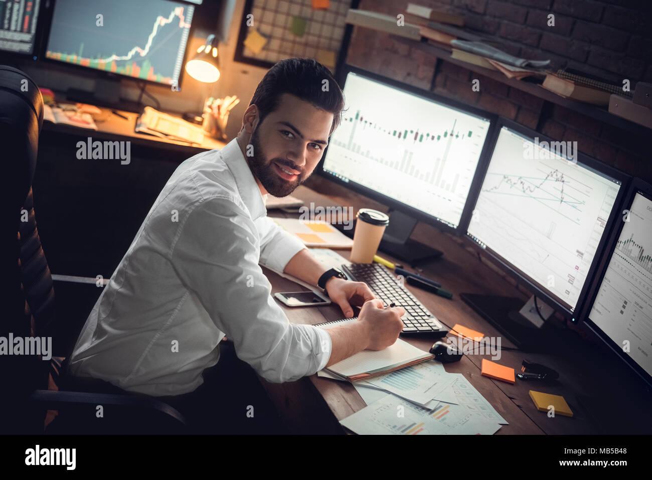 Young bearded man trader at office sitting at table looking camera smiling happy working Stock Photo