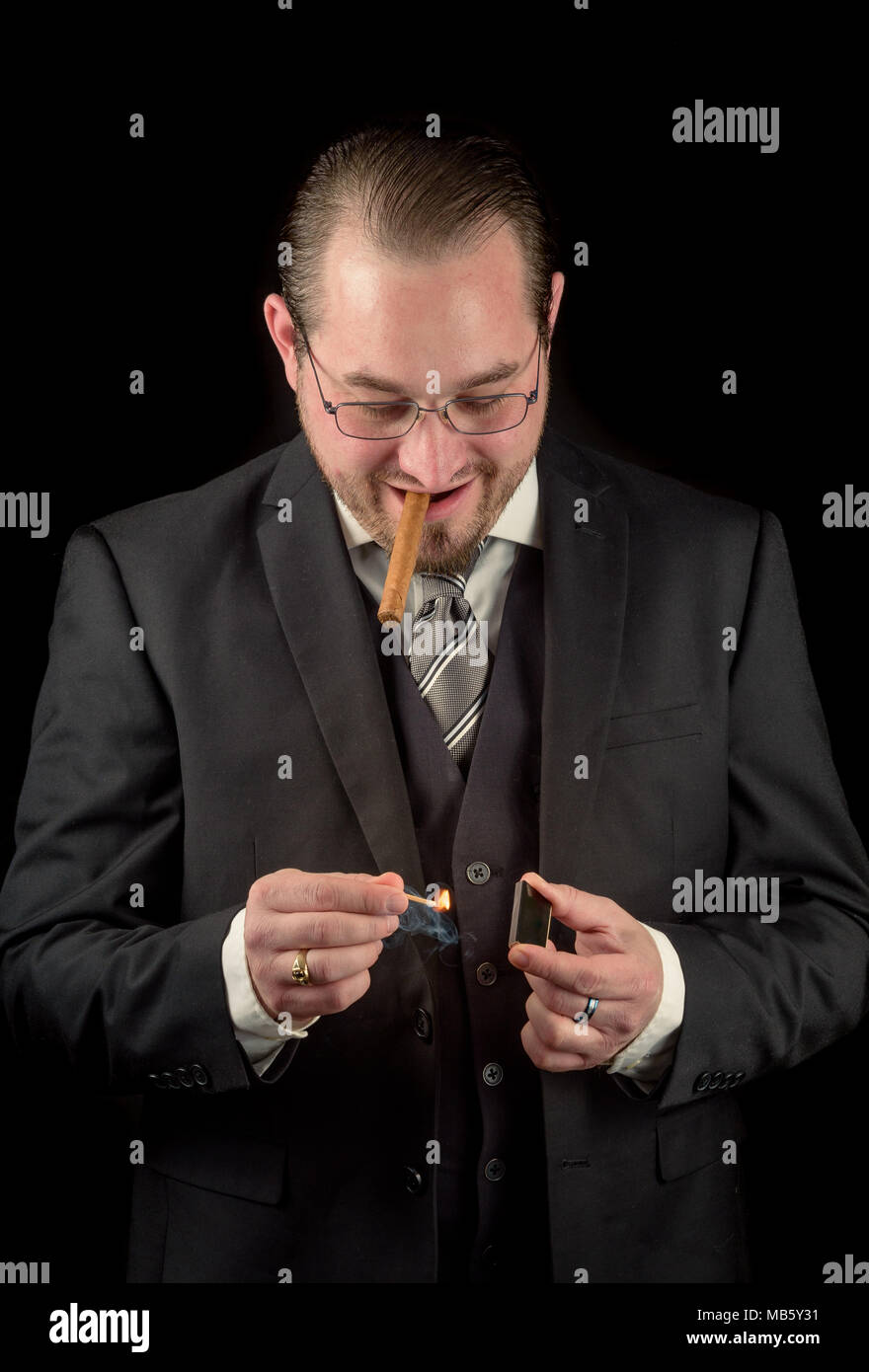 Caucasian male model with glasses in a black suit isolated against a black backgroud. Model striking a match with a fresh cigar in mouth Stock Photo