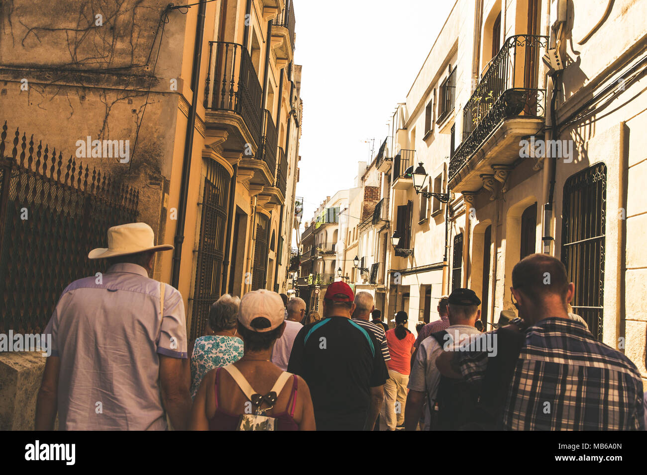 Tourists walking down a street in Spain. Some tourists are wearing caps, an elderly man wears a hat. Stock Photo