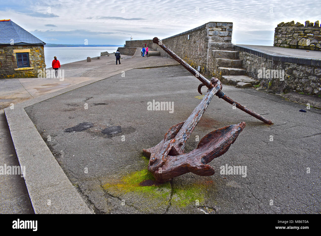 Old anchor with people strolling behind the high solid stone wall which forms the Porthcawl Harbour breakwater with the iconic lighthouse in distance. Stock Photo