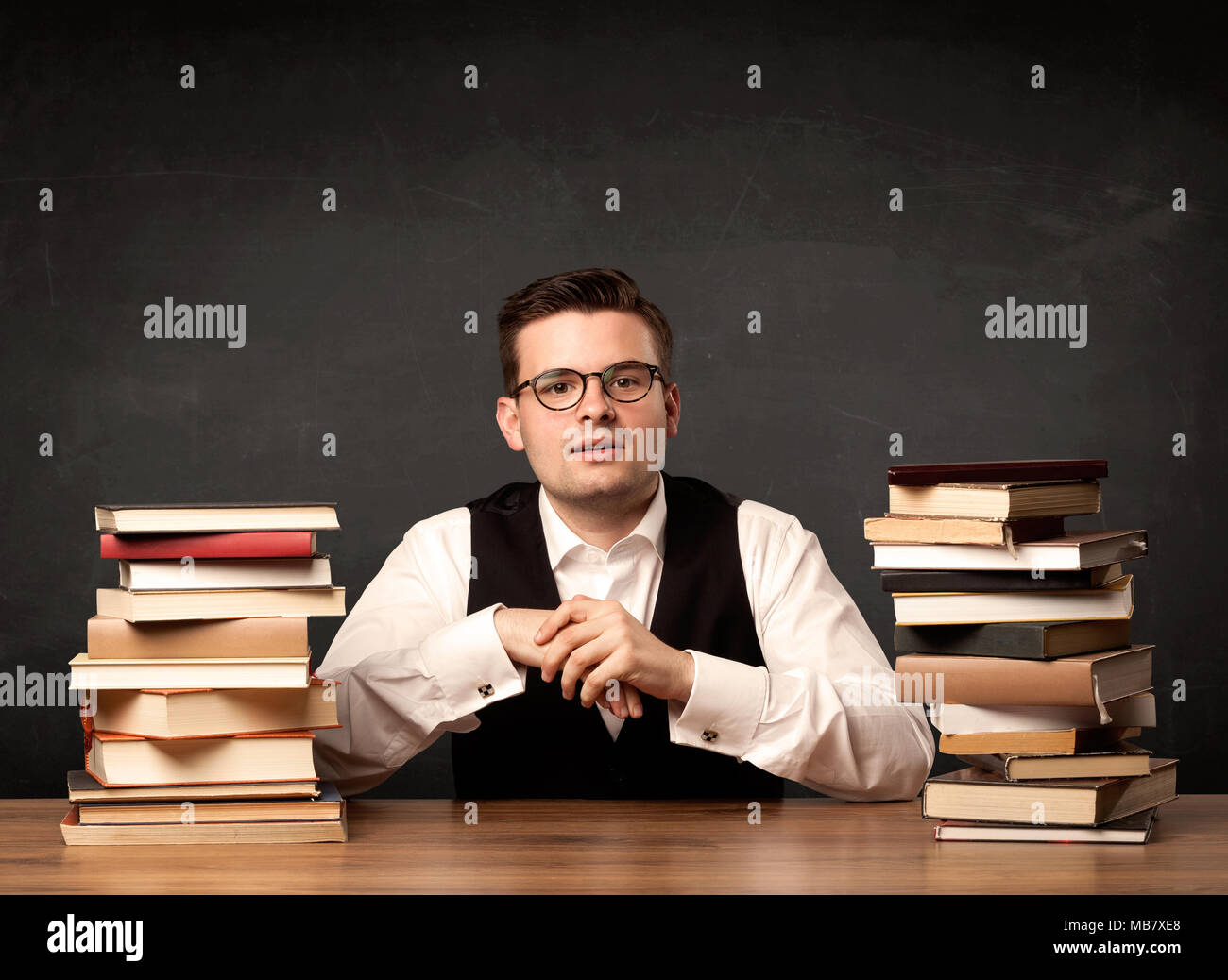 A young teacher in glasses sitting at classroom desk with pile of books ...