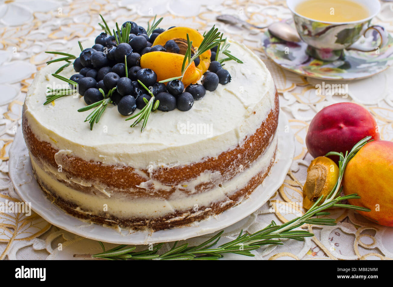 side view of delicious homemade cake with cream, berries, rosemary and nectarine on top on a decorative white table cloth next to a vintage cup of tea Stock Photo