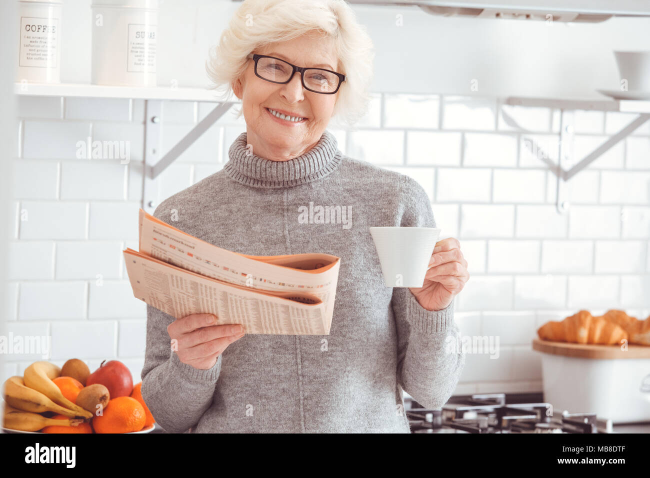 Businesswoman read newspaper, drink tea looking at camera, toothy smiling. Indoor, studio shoot, kitchen interior Stock Photo