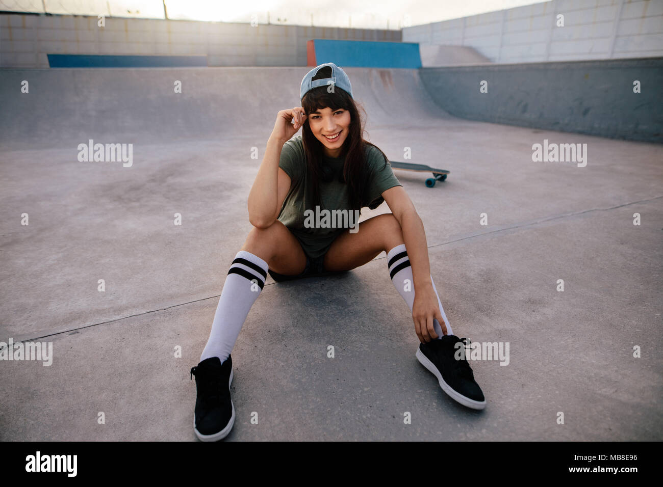 Portrait of attractive urban girl sitting relaxed at skate park. Female skateboarder taking break after skate boarding outdoors. Stock Photo