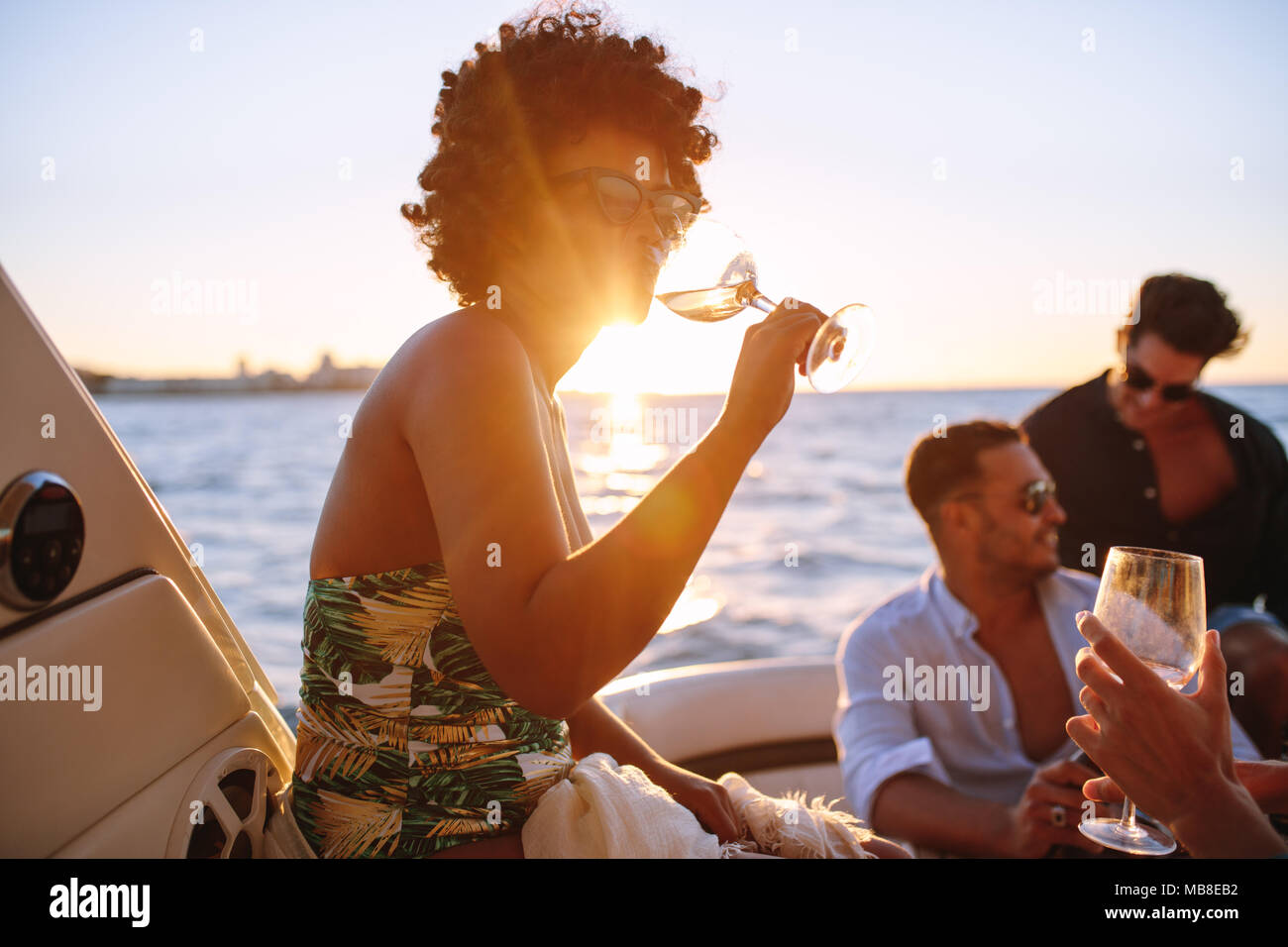 African woman drinking during sunset boat party with friends. Group of men and women having a boat party during sunset. Stock Photo