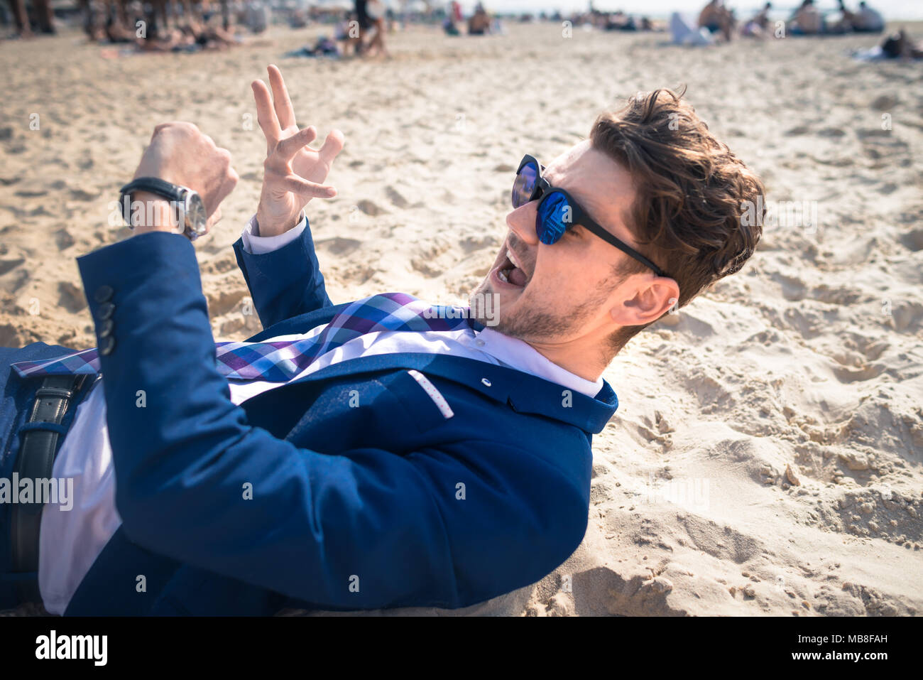 Freaky young man in elegant suit is late on the work. Stylish guy active gesticulate and looks on the wristwatch on the beach. Funny manager Stock Photo