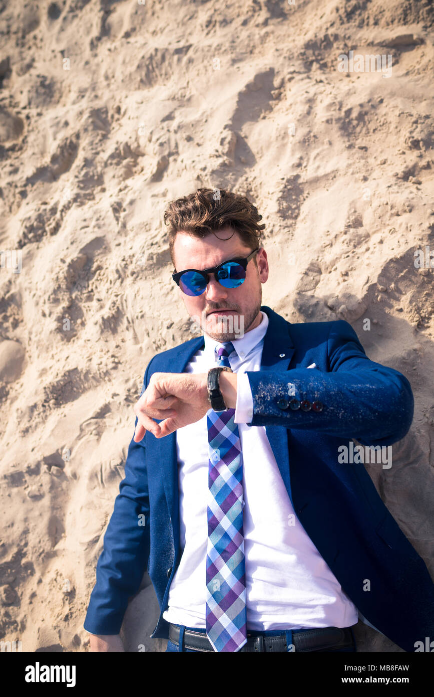 Funny manager lost in time but stays calm. Serious young man in elegant suit is late on the work. Stylish guy lies on the beach and looks on the watch Stock Photo