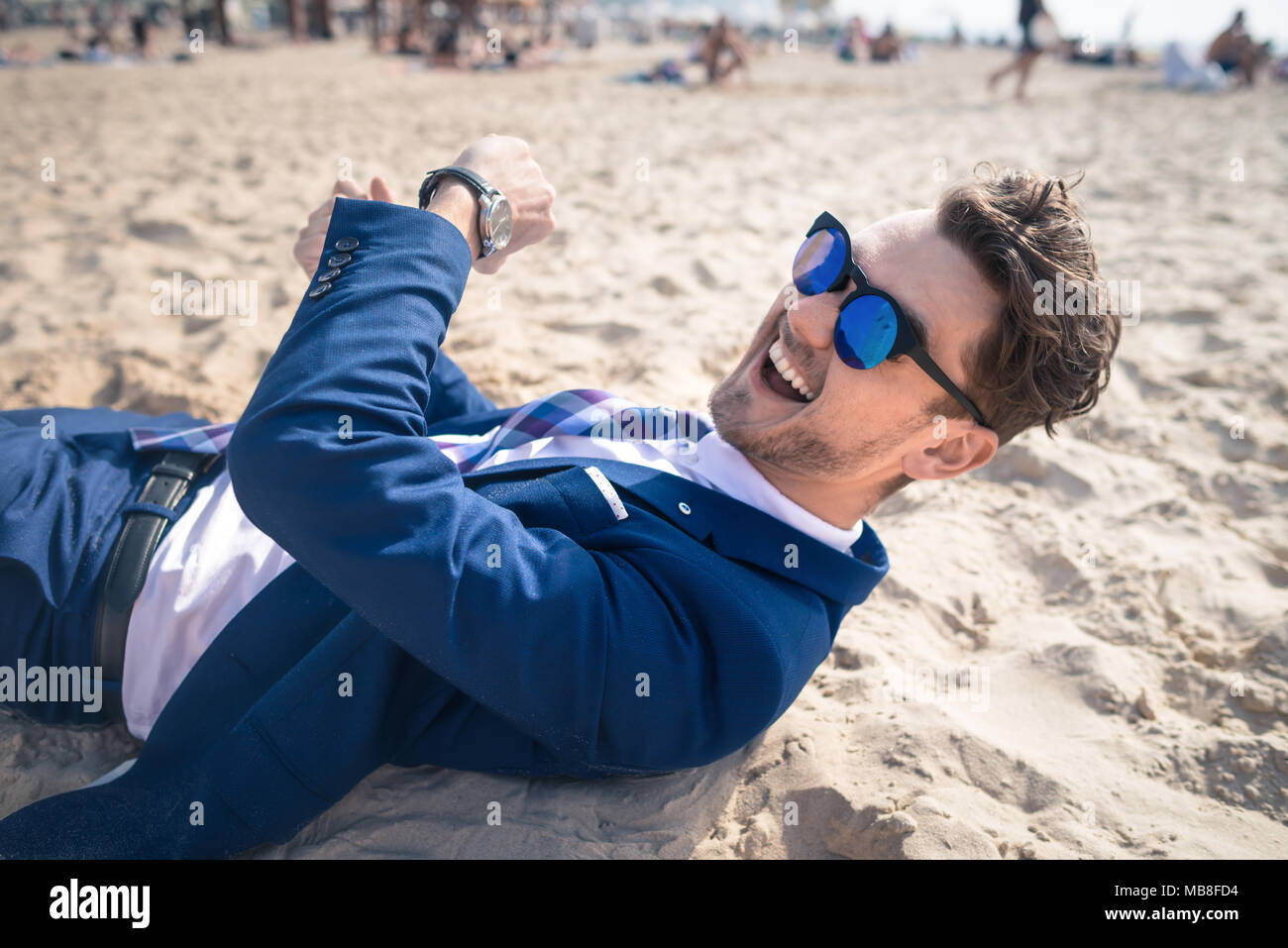 Unusual young man in elegant suit relaxes on the beach and laughs. Funny guy lies down on the sand and has time to rest Stock Photo