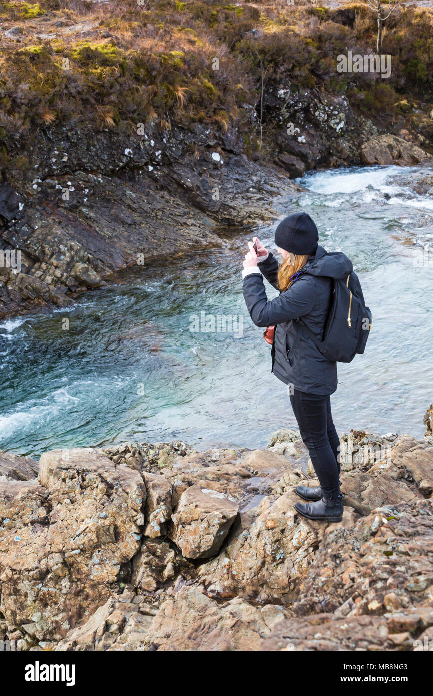 Young woman taking a photo with mobile phone standing on rocks at Fairy Pools, river Brittle, Isle of Skye, Scotland, UK in March Stock Photo