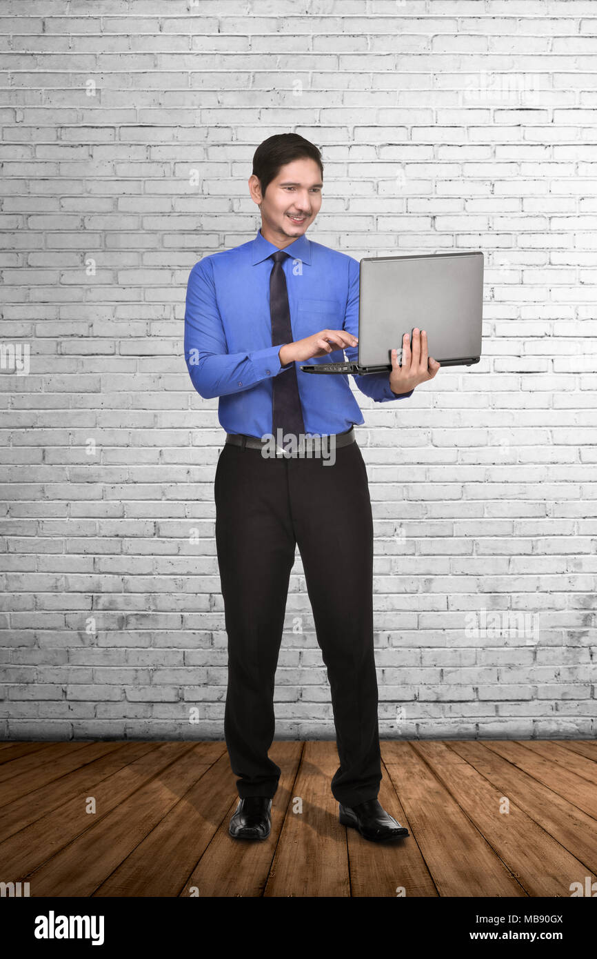 Young asian businessman standing while typing on laptop with white wall ...