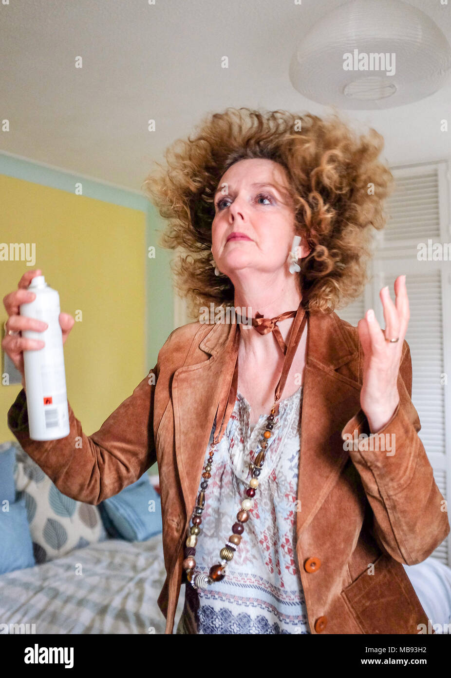 Middle aged mature woman using hair spray on highlighted blonde curly hair ready for ash evening out Photograph taken by Simon Dack Stock Photo