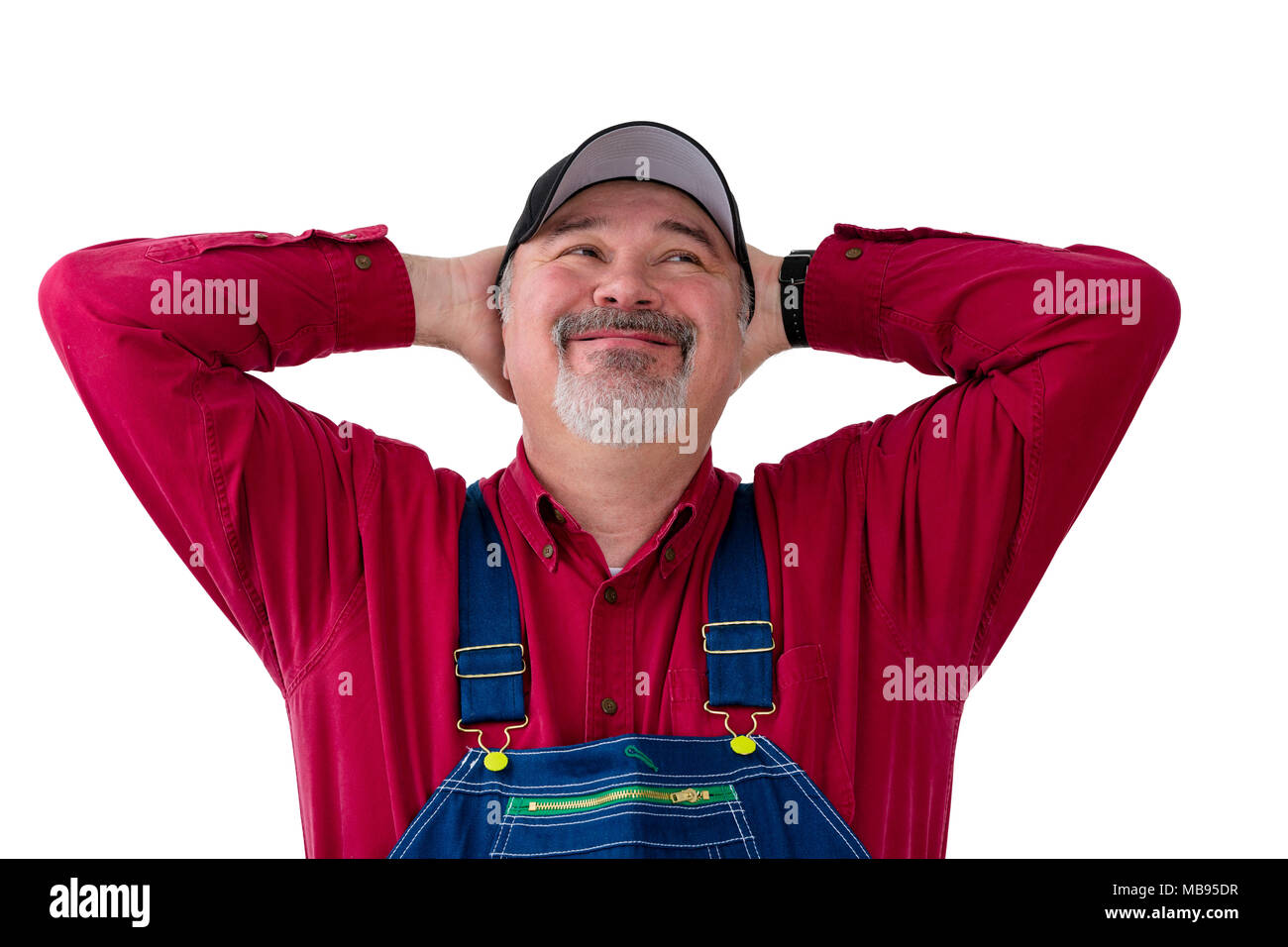 Farmer or worker in overalls and cap standing with his hands behind his head looking up with a big happy smile of satisfaction over white Stock Photo
