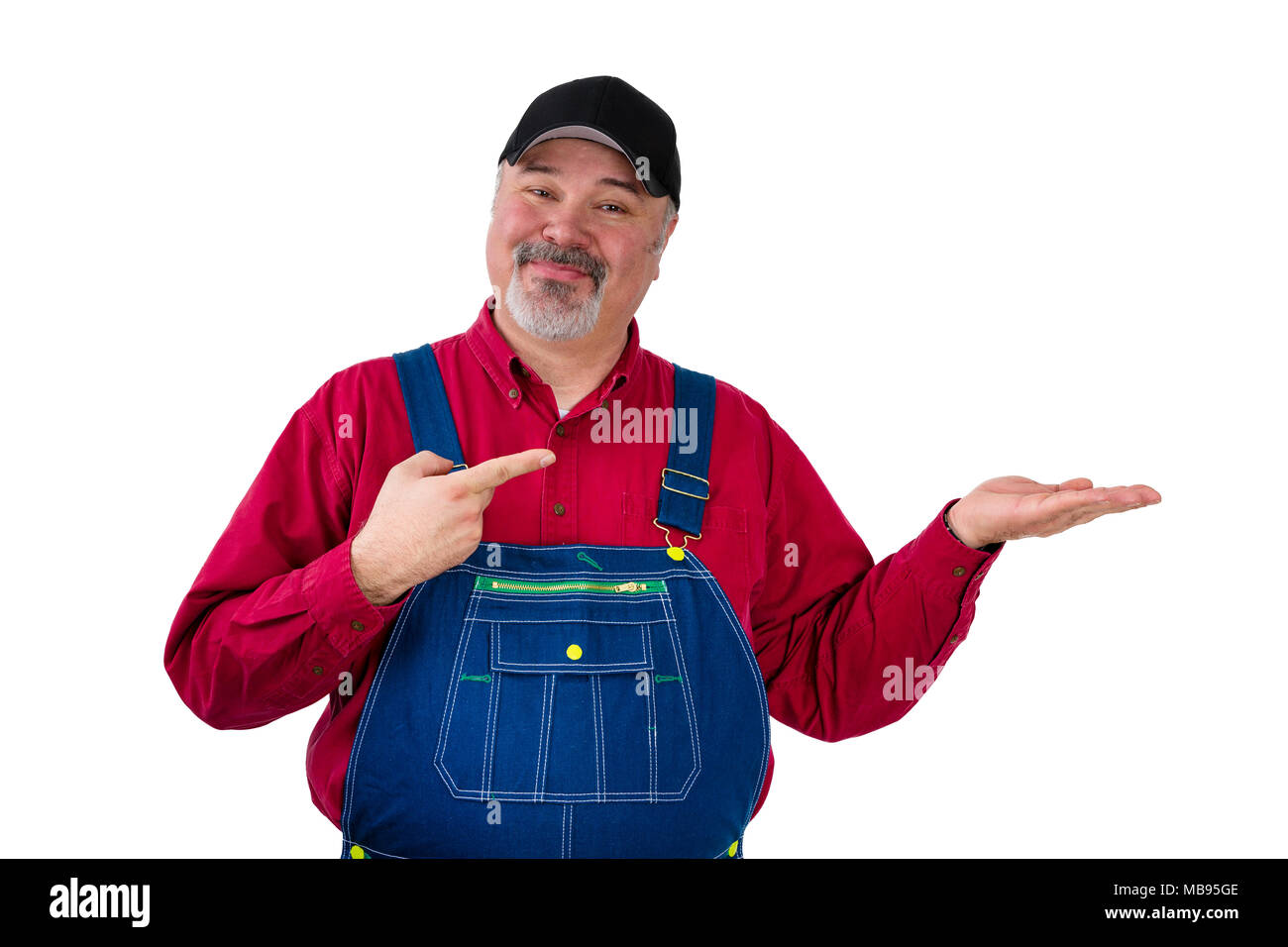 Smiling bearded worker in cap and bib overall holding out his empty hand over blank white copy space pointing to it with a smile of satisfaction Stock Photo