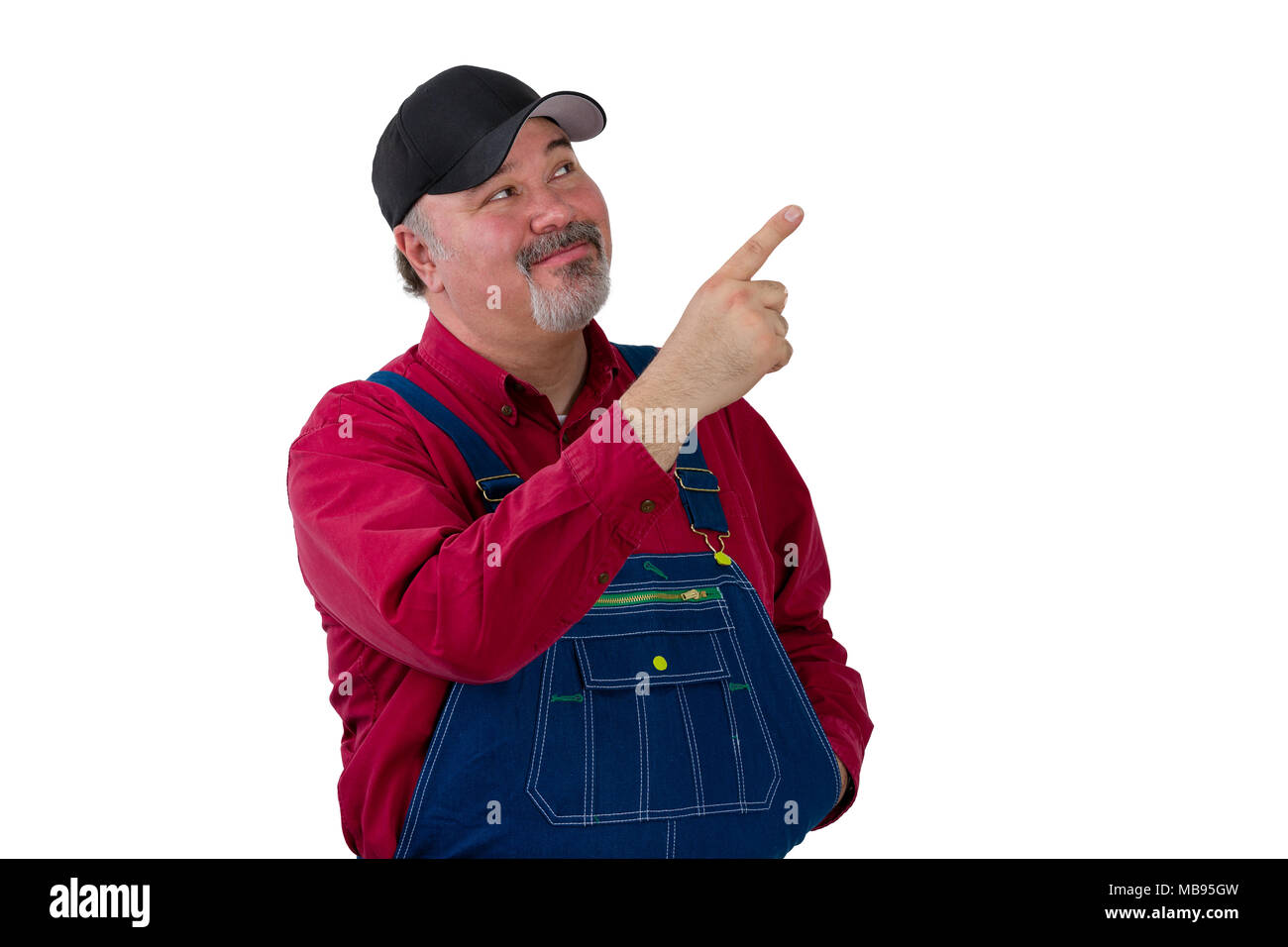 Smiling middle-aged worker in dungarees and cap pointing upwards towards blank copy space on white Stock Photo