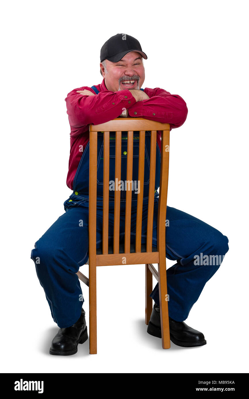Jovial vivacious worker in denim overalls sitting on a reversed wooden chair leaning on the back laughing over a white background Stock Photo