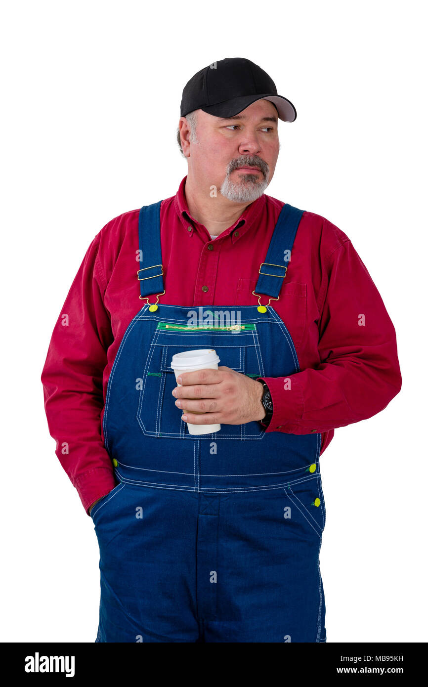 Thoughtful worker or farmer in denim dungarees and cap standing watching to the side with a serious contemplative expression while holding coffee isol Stock Photo