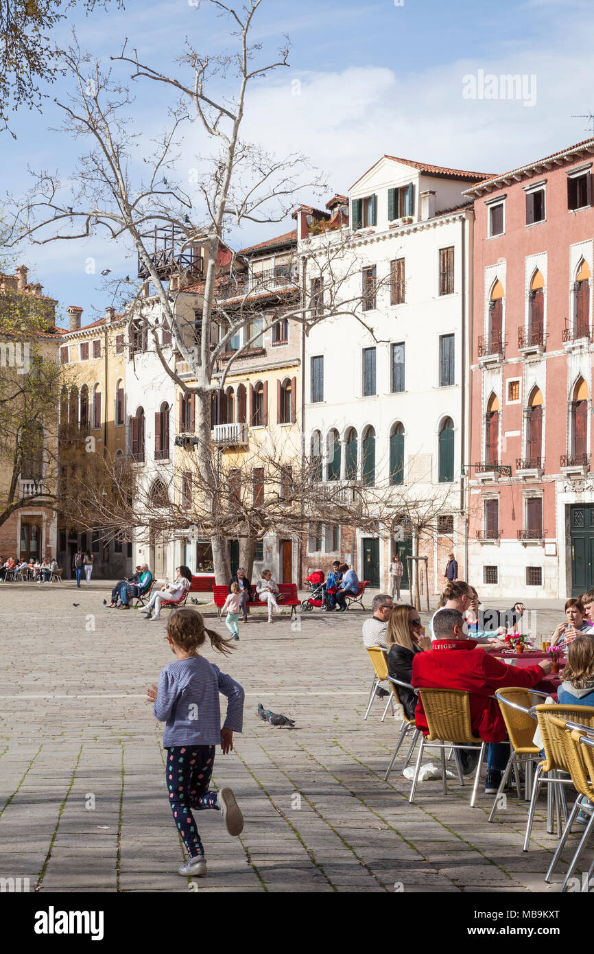 Campo San Polo, San Polo, Venice, Veneto, Italy in spring with a little girl chasing birds and people dining at a restaurant  and relaxing on benches  Stock Photo
