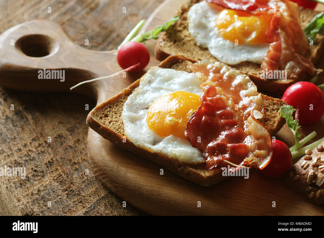 Breakfast of crispy bacon, fried eggs and bread. Sandwiches on cutting board. Rustic table Stock Photo