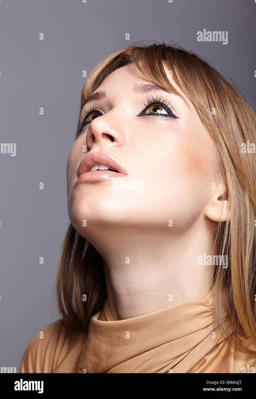 Portrait of blonde young woman looking up. Female with green eyes and long straight hair. Girl dressed in beige dress on gray background. Stock Photo