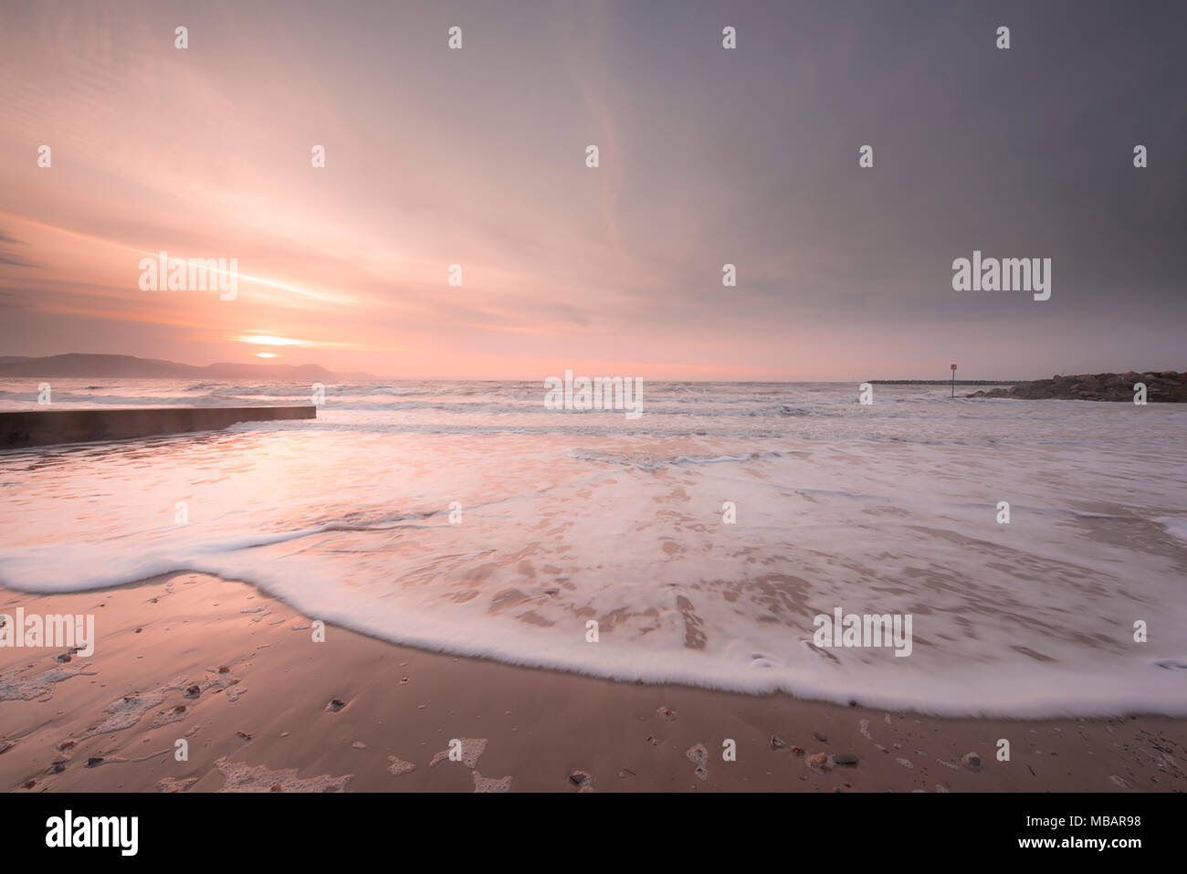 Beautiful pale pink tones in the sky and sea at sunrise as morning waves  gently roll onto the sandy beach in Lyme Regis. Stock Photo