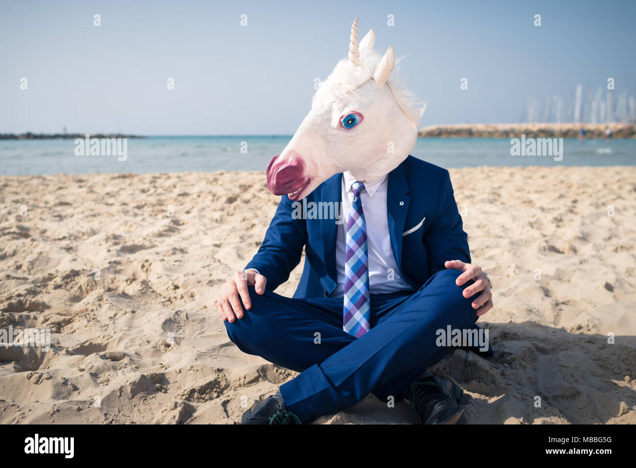Young trendy man in funny mask and elegant suit on vacation. Unicorn sits on the beach on background of sea and sky Stock Photo