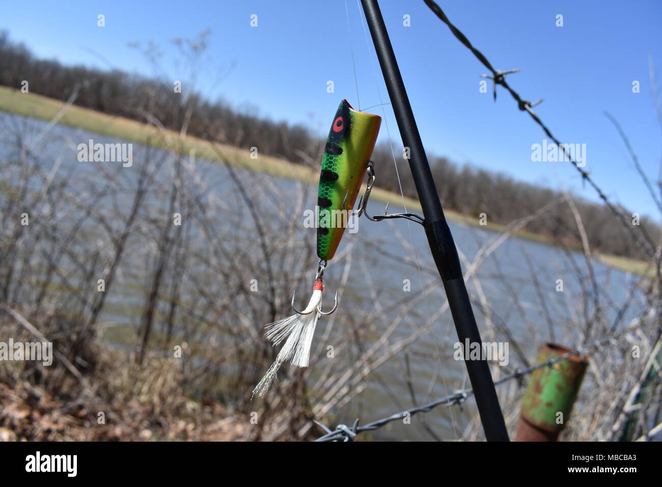 A close-up of a bass lure dangling from a fishing pole and a pond in the background calls to the fisherman. Rural Missouri, MO, United States, USA, US Stock Photo