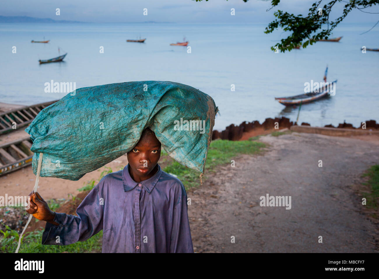 YONGORO, SIERRA LEONE - June 05, 2013: West Africa, unknown boy carries a bag on his shoulders near the beach with fishing boats in front of the capit Stock Photo