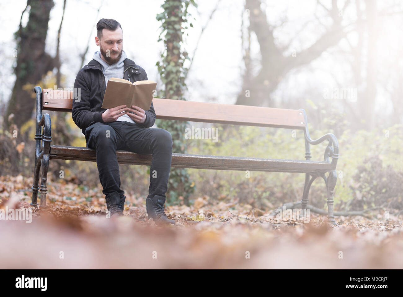A handsome young hipster man sitting on a bench and reading a book in ...