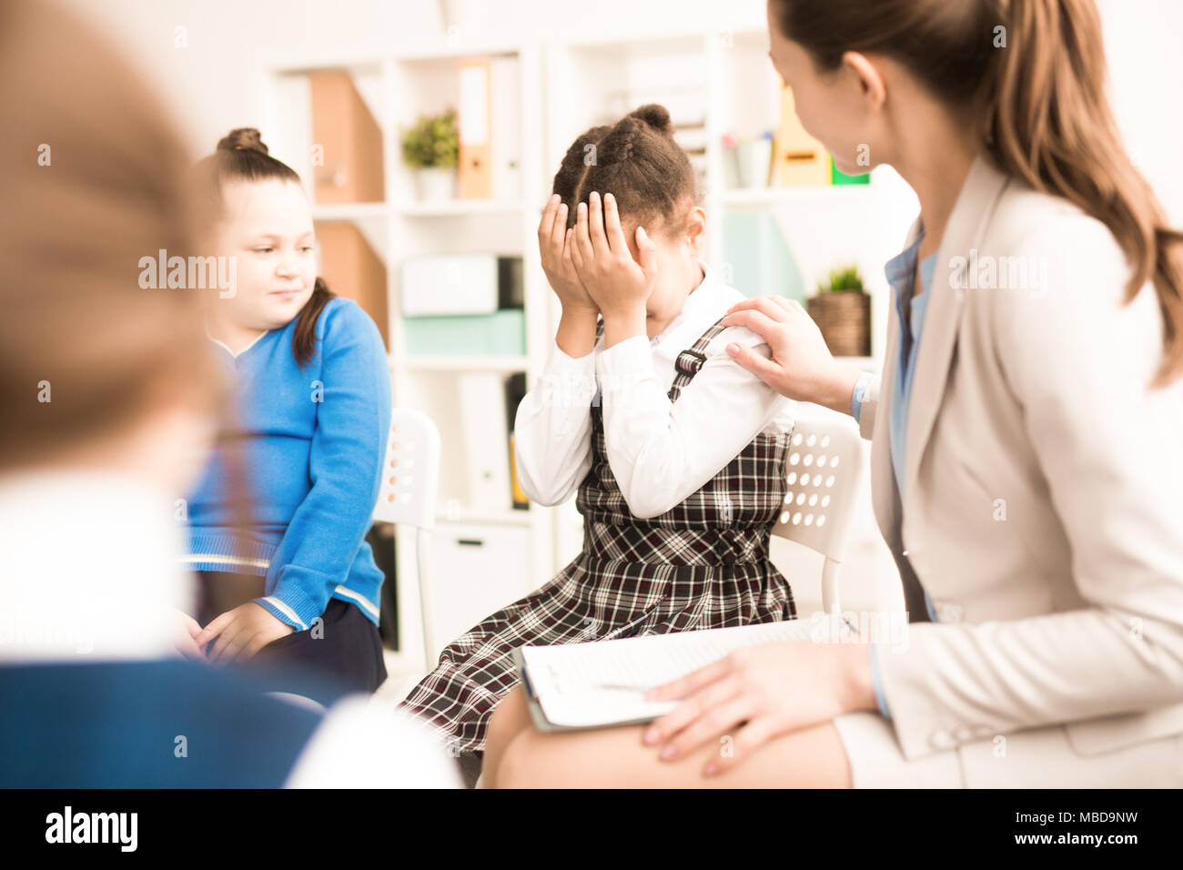 Crying girl sitting on chair nearby with school psychologist or teacher trying to comfort her Stock Photo