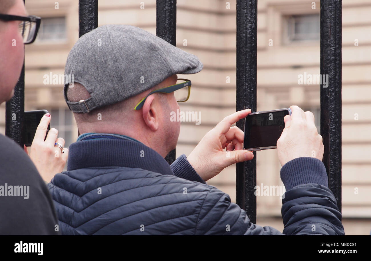 A man wearing a flat cap taking a photograph of Buckingham Palace through the railings with his smartphone Stock Photo