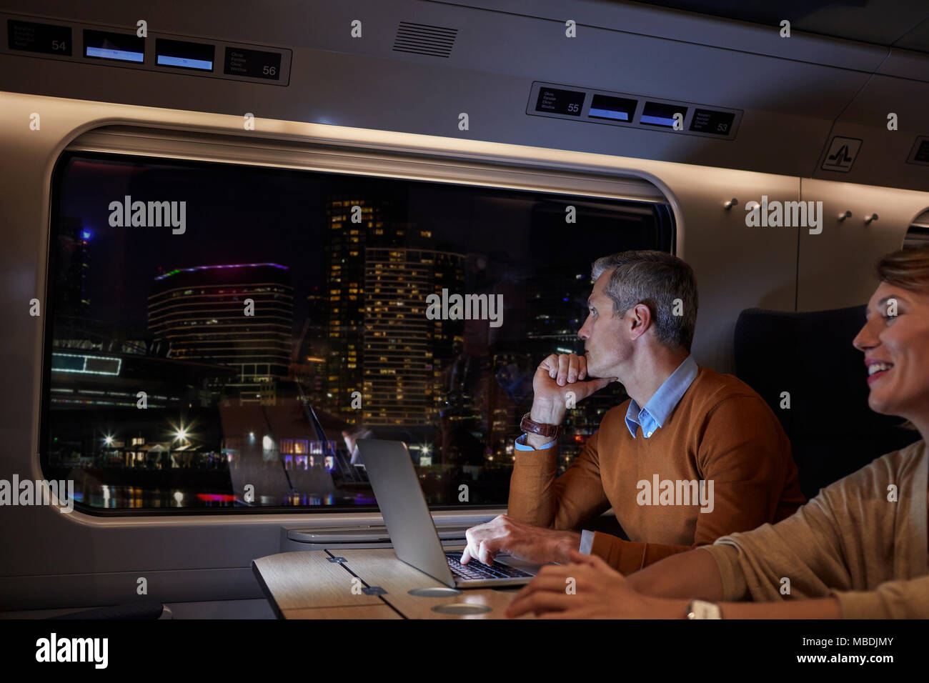 Businessman working at laptop on passenger train at night, looking out window at passing city Stock Photo