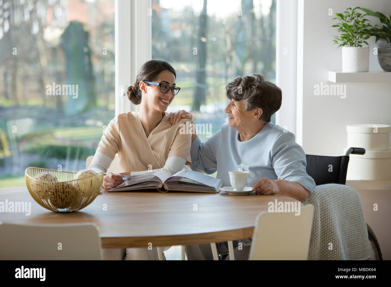 Happy pensioner and nurse sitting together at the table and having a conversation Stock Photo