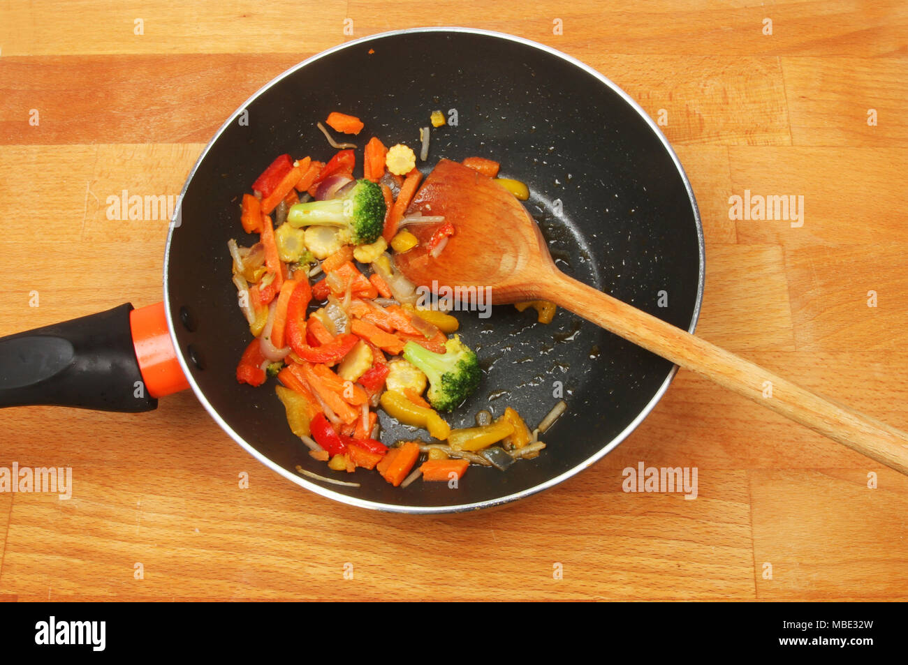 Stir fried vegetables in a wok with a wooden spoon on a wooden worktop Stock Photo