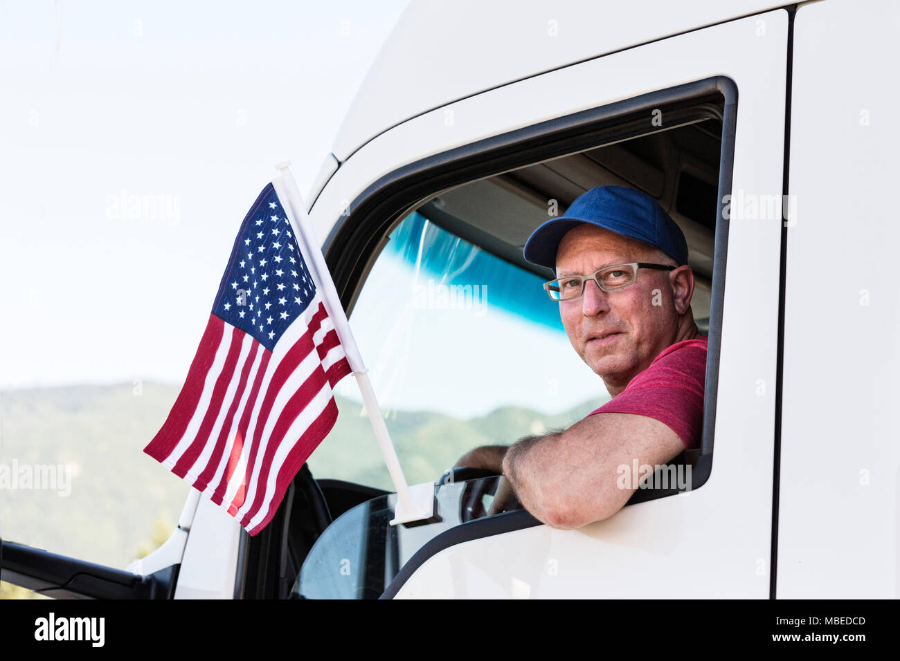 Caucasian man truck driver in the cab of his truck with an American Flag attached to the window. Stock Photo