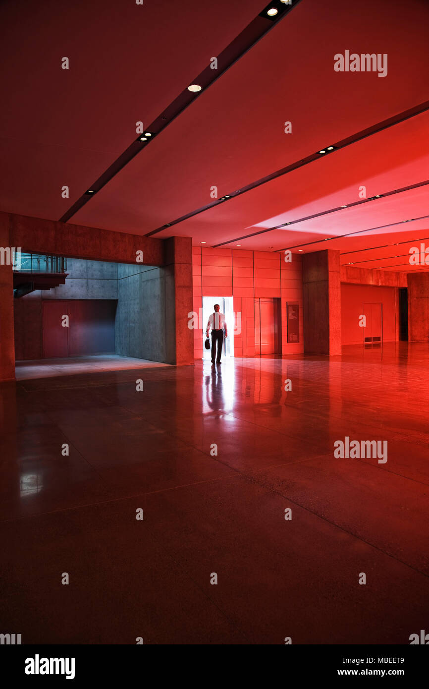 Businessman walks through the door of a lobby area that is lit by daylight through red tinted glass. Stock Photo