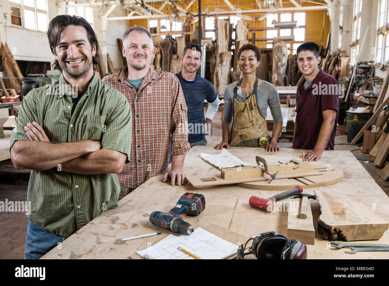 Team of multi-racial factory workers standing next to a work station in a large wood working factory. Stock Photo