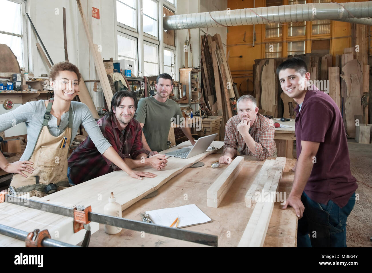 Team of multi-racial factory workers standing next to a work station in a large wood working factory. Stock Photo