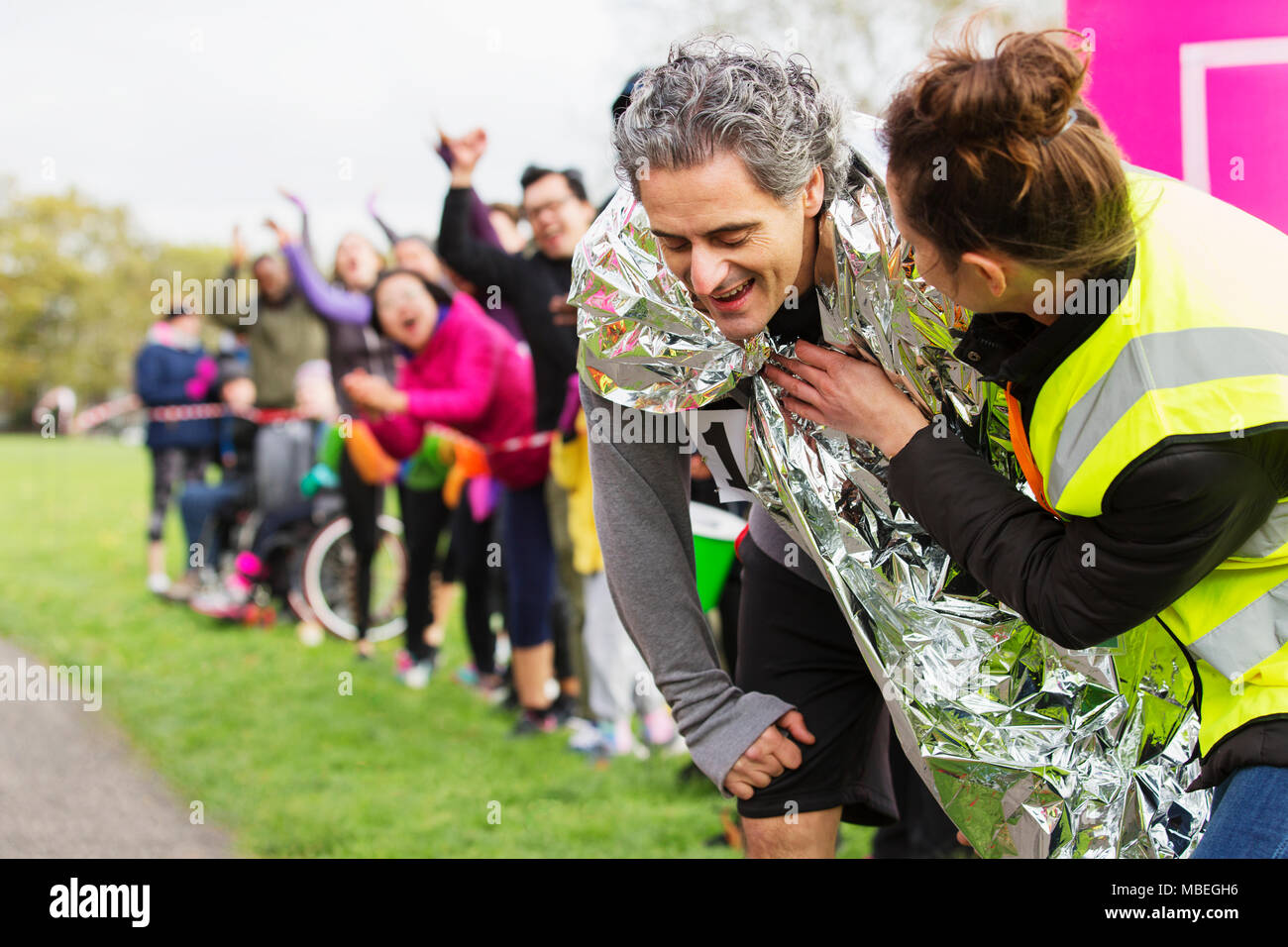 Woman wrapping thermal blanket around exhausted male runner finishing marathon Stock Photo