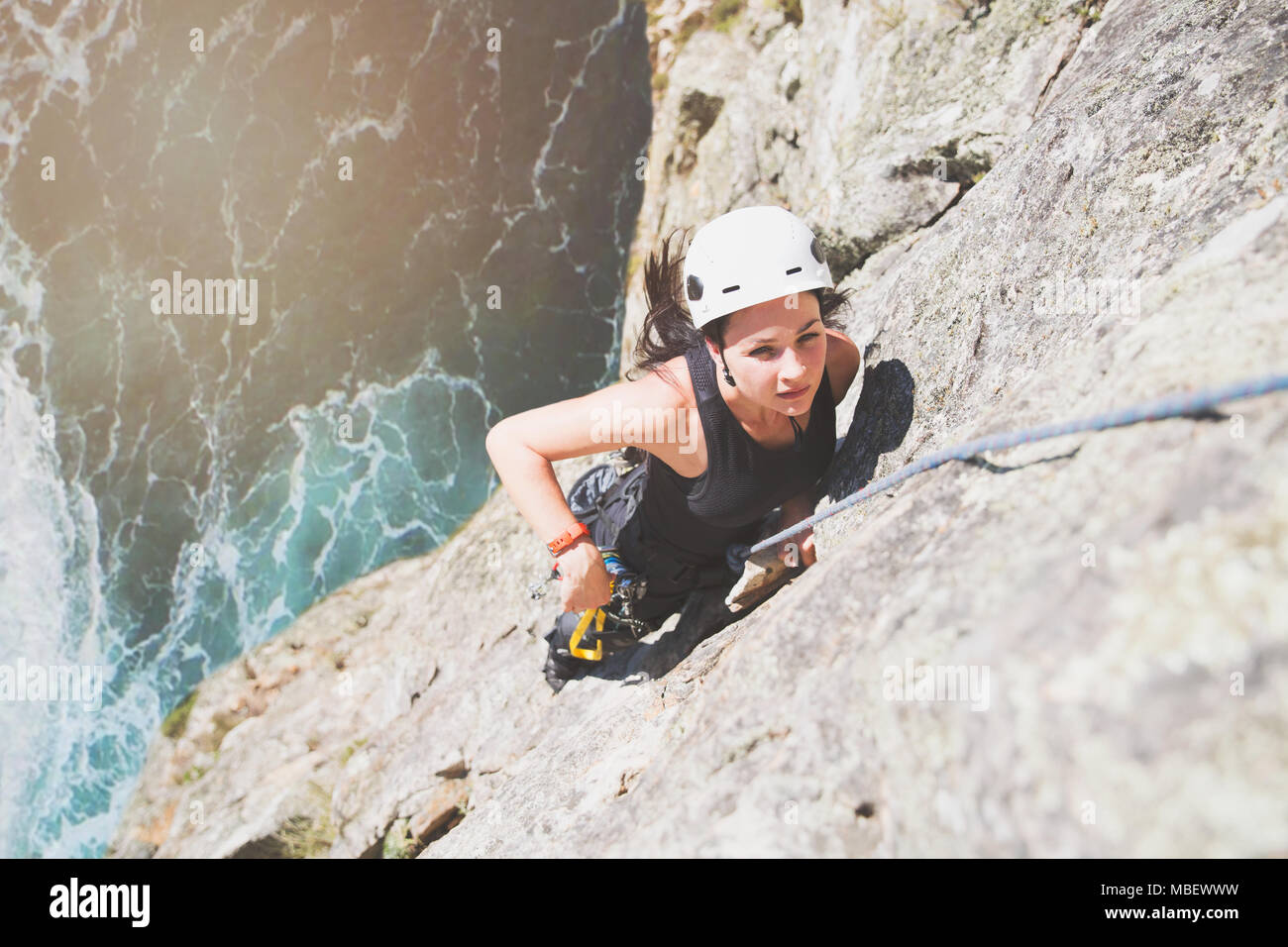 Focused, determined female rock climber scaling rock above sunny ocean Stock Photo