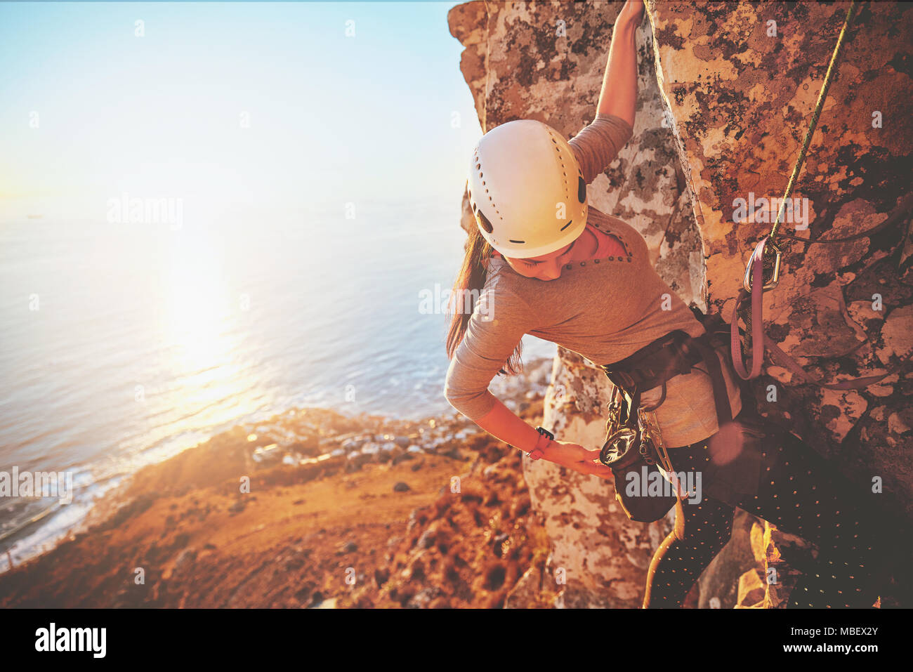 Female rock climber reaching for clip above sunny ocean Stock Photo