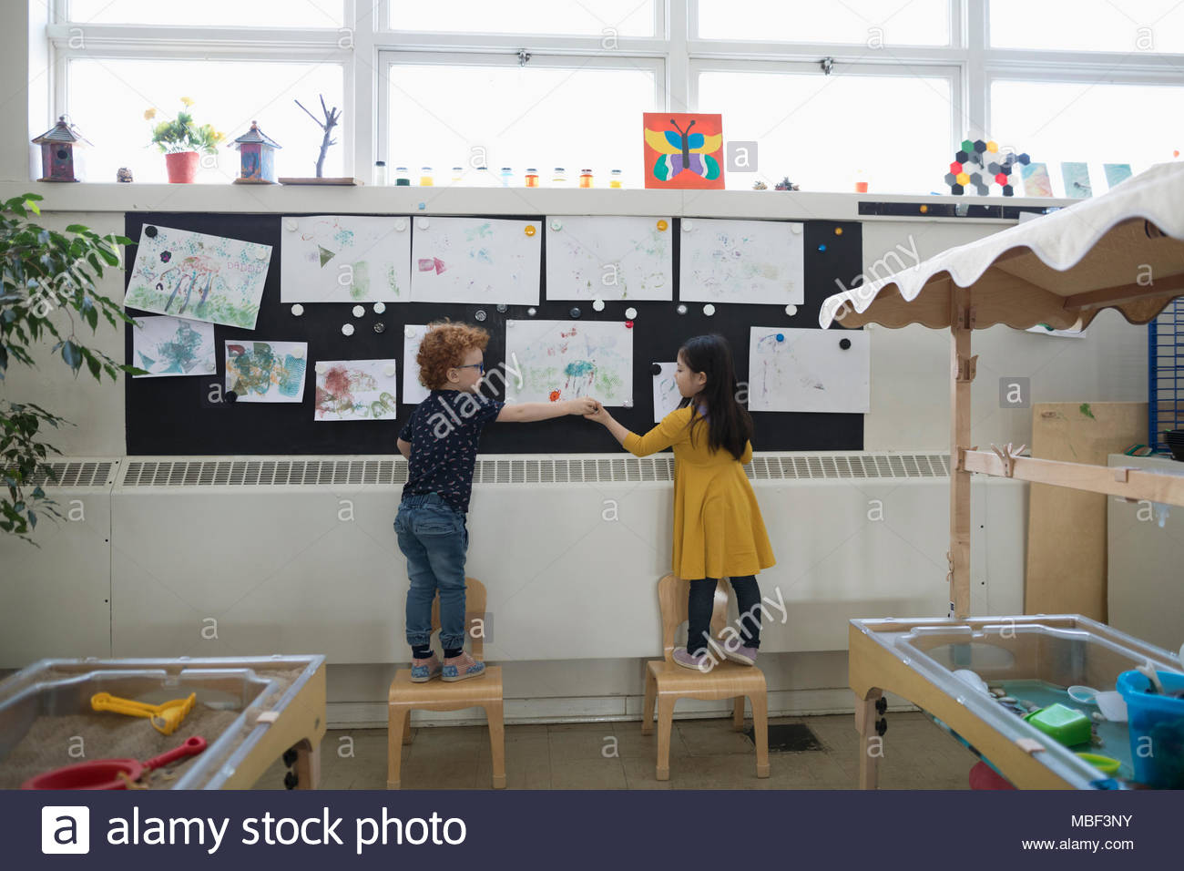 Preschool boy and girl hanging paintings on wall in classroom Stock