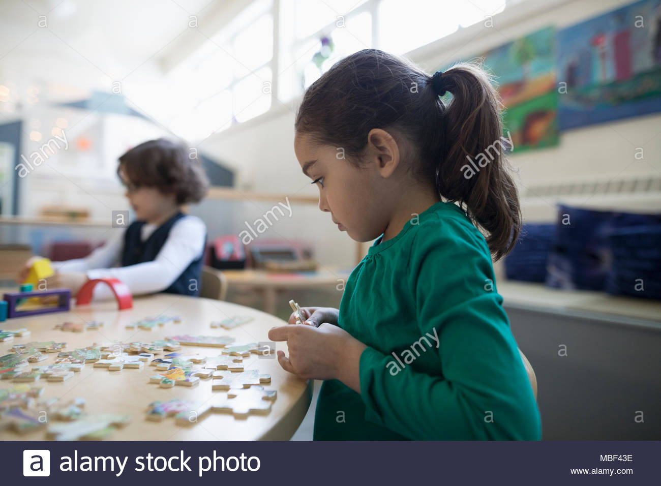 Focused preschool girl assembling jigsaw puzzle in classroom Stock