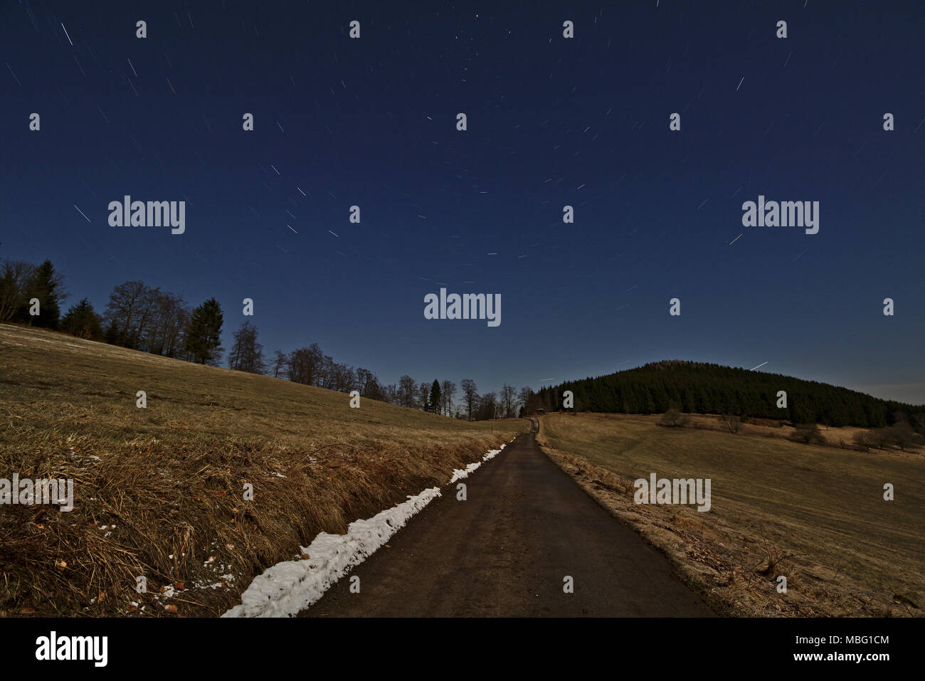 Starry night sky with star trails rotating around the North Star above a single lane road leading through fields Stock Photo